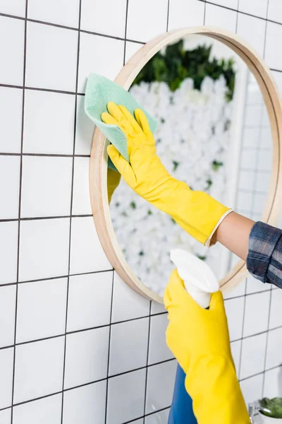 Cropped view of woman in yellow rubber gloves cleaning mirror with detergent and rag in bathroom — Stock Photo