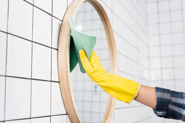 Обрезанный вид женщины в резиновых перчатках с зеркалом с тряпкой во время уборки ванной комнаты — стоковое фото