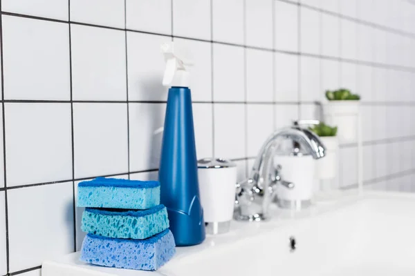 Blue sponges and bottle of detergent on sink in bathroom on blurred background — Stock Photo