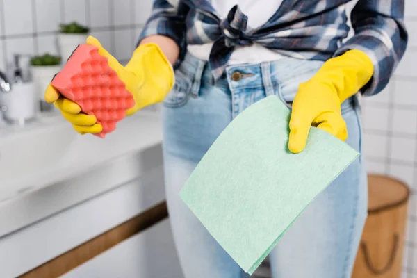 Vista recortada de trapo y esponja en manos de mujer en guantes de goma sobre fondo borroso en el baño - foto de stock