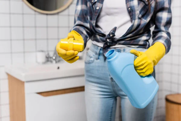 Cropped view of bottle of cleaner and cap in hands of woman in rubber gloves on blurred background in bathroom — Stock Photo