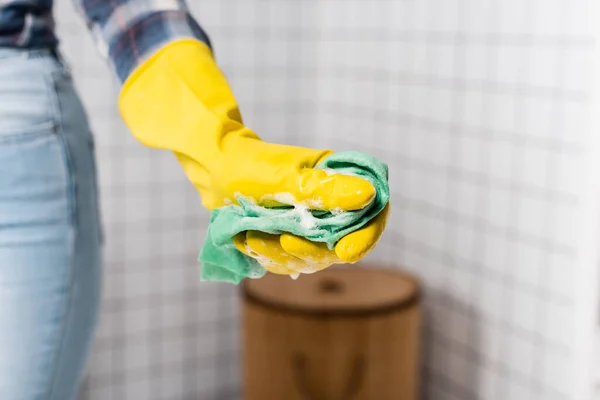 Cropped view of rag with soap in hand of woman in rubber glove on blurred background in bathroom — Stock Photo