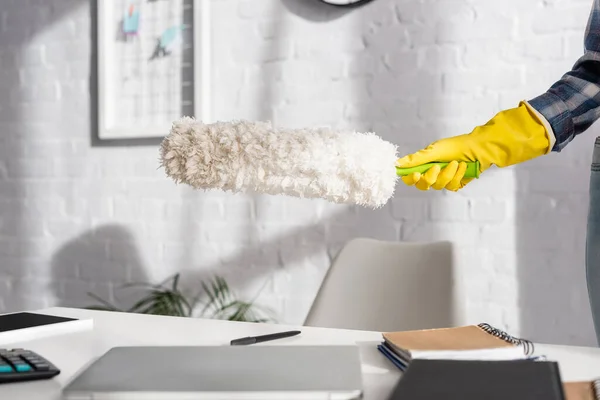 Vista cortada da mulher em luva de borracha segurando escova de poeira perto de dispositivos e notebook em primeiro plano borrado na mesa — Fotografia de Stock