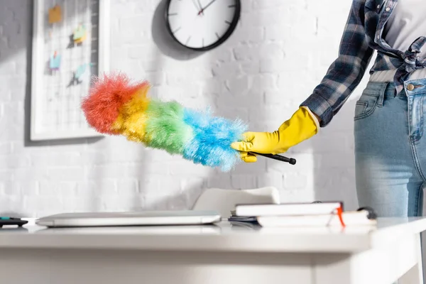 Cropped view of woman in rubber glove holding dust brush near laptop an notebooks on blurred foreground — Stock Photo