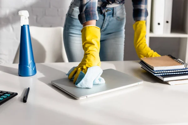 Ausgeschnittene Ansicht einer Frau in Gummihandschuhen, die Laptop in der Nähe von Waschmittel und Notizbücher auf verschwommenem Hintergrund auf dem Tisch säubert — Stockfoto