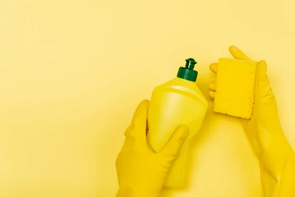 Vue du dessus des mains en gants de caoutchouc tenant une bouteille de liquide vaisselle et une éponge sur fond jaune — Photo de stock