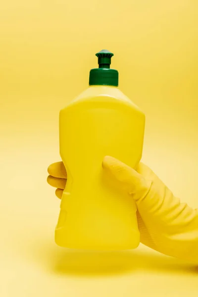 Vue recadrée de la main dans un gant en caoutchouc tenant une bouteille de liquide vaisselle sur fond jaune — Photo de stock