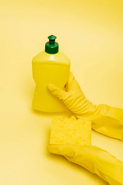 Vista en ángulo alto de las manos con guantes de goma que sujetan la esponja y el líquido lavavajillas sobre fondo amarillo - foto de stock