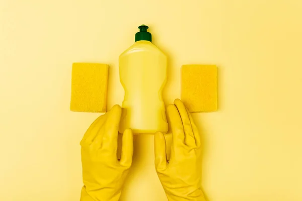 Vista dall'alto di persona in guanti di gomma che tiene il liquido di lavaggio dei piatti vicino a spugne su sfondo giallo — Foto stock