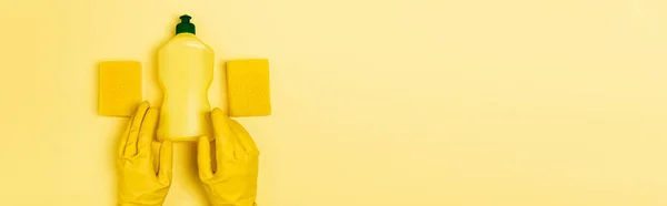 Vue du dessus du liquide vaisselle et des éponges près des mains dans des gants en caoutchouc sur fond jaune, bannière — Photo de stock