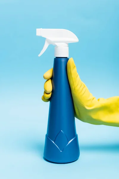 Main en gants de caoutchouc jaune tenant détergent avec pulvérisateur sur fond bleu — Photo de stock