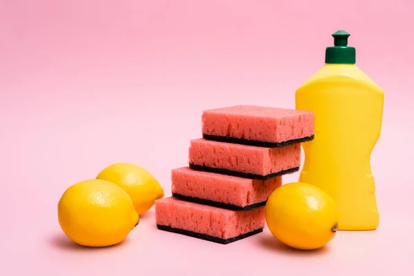 Limones frescos cerca de esponjas y líquido lavavajillas sobre fondo rosa - foto de stock