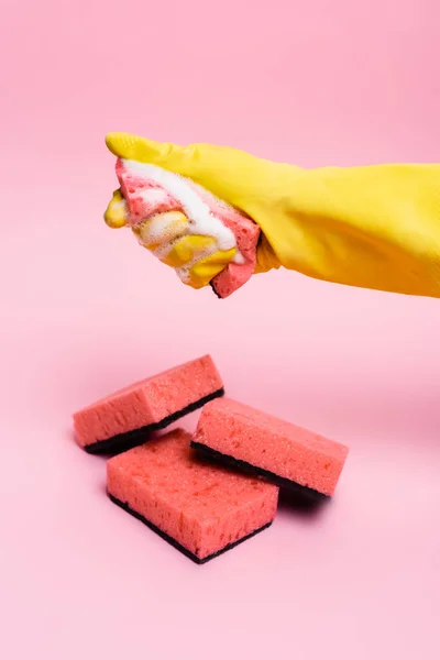 Hand in Gummihandschuh hält Schwamm mit Seifenlaugen in der Nähe von Schwämmen auf rosa Hintergrund — Stockfoto