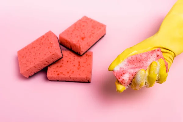 Vue du dessus de la main en gant frotté tenant éponge avec des savons près des éponges floues sur fond rose — Photo de stock