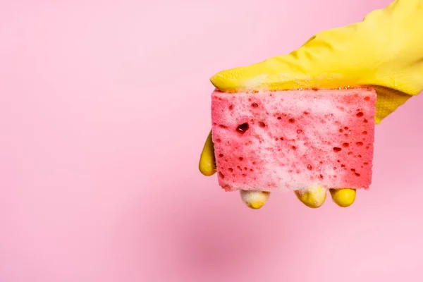 Vista recortada de la mano en guante frotado sosteniendo esponja con espuma de jabón sobre fondo rosa - foto de stock