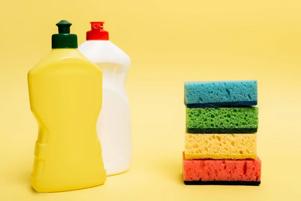 Botellas de líquido lavavajillas y esponjas de colores sobre fondo amarillo - foto de stock