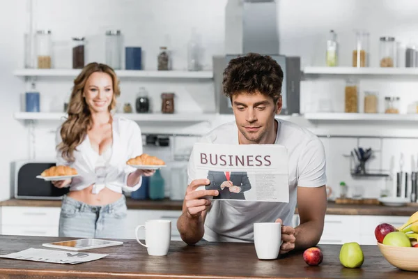 Man reading business newspaper near smiling girlfriend with croissants on blurred background — Stock Photo