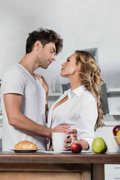 Sexy pareja abrazándose y mirándose el uno al otro cerca del desayuno en primer plano borroso en la mesa - foto de stock