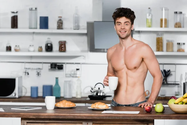 Homem muscular sorrindo para a câmera enquanto segurando copo perto do café da manhã na mesa da cozinha — Fotografia de Stock