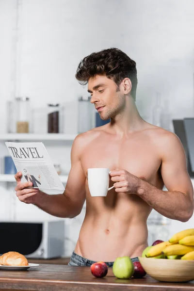 Uomo muscolare leggere notizie di viaggio e tenere la tazza durante la colazione in cucina — Foto stock