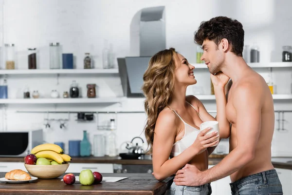 Улыбающаяся женщина в лифчике держит чашку и трогает сексуального парня на кухне — стоковое фото
