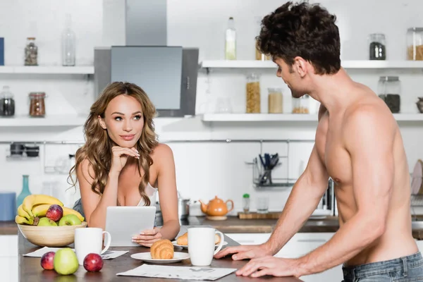 Сексуальная женщина с цифровым планшетом смотрит на парня без рубашки рядом с завтраком на размытом переднем плане на кухне — стоковое фото