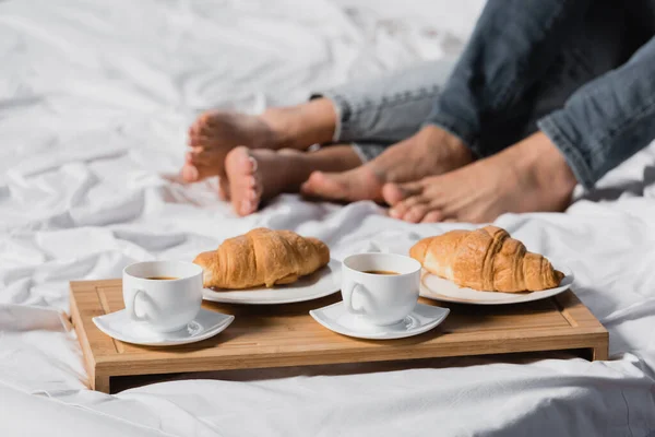 Vista coping de café e croissants na bandeja de café da manhã perto das pernas do casal no fundo embaçado na cama — Fotografia de Stock