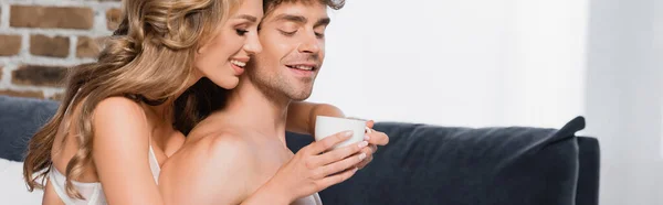 Femme souriante en soutien-gorge tenant tasse de café et étreignant homme torse nu à la maison, bannière — Photo de stock