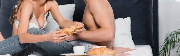 Обрізаний вид сексуальної жінки, що тримає круасан біля чашки кави і без сорочки хлопець на ліжку, банер — стокове фото