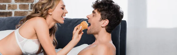 Donna positiva in reggiseno alimentazione fidanzato con croissant sul letto, banner — Foto stock
