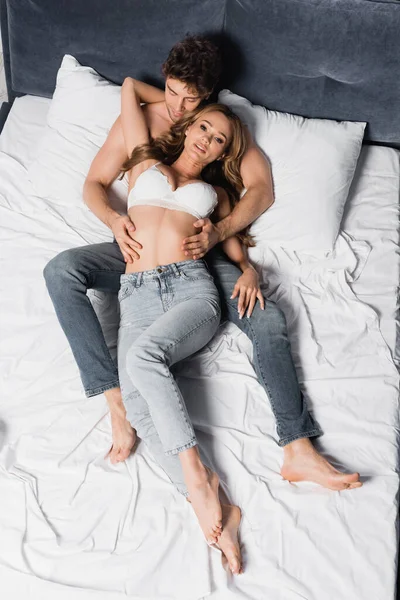 Top view of shirtless man in jeans touching seductive woman looking at camera on bed — Stock Photo