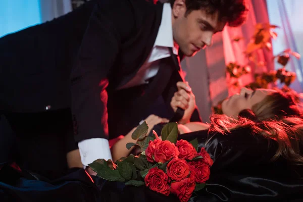 Roses near sexy woman holding tie of boyfriend on blurred background on bed — Stock Photo