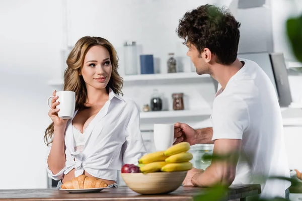 Femme sexy tenant tasse près du petit déjeuner et petit ami sur le premier plan flou dans la cuisine — Photo de stock