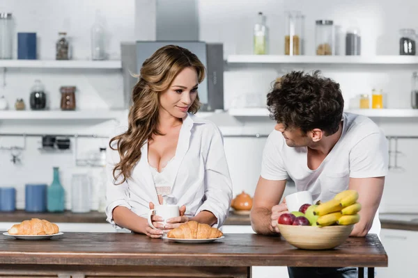 Mujer sexy sosteniendo la taza y mirando al novio cerca de croissants y frutas en primer plano borrosa - foto de stock