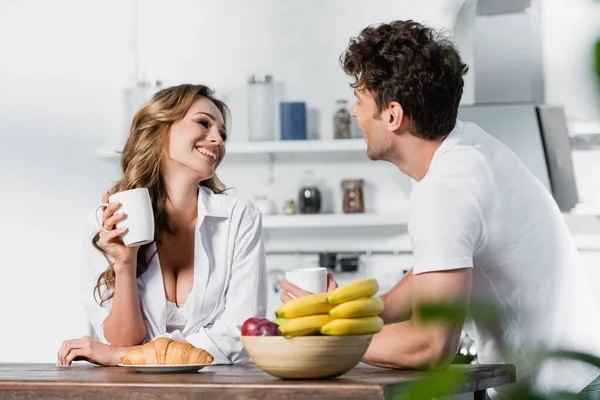 Smiling woman in shirt and bra holding cup near croissant, fruits and boyfriend on blurred foreground — Stock Photo