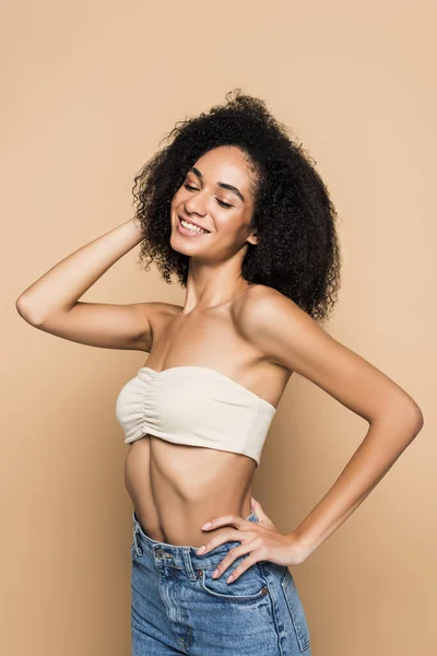 Cheerful african american woman with bare shoulders in top and jeans standing with hand on hip on beige — Stock Photo
