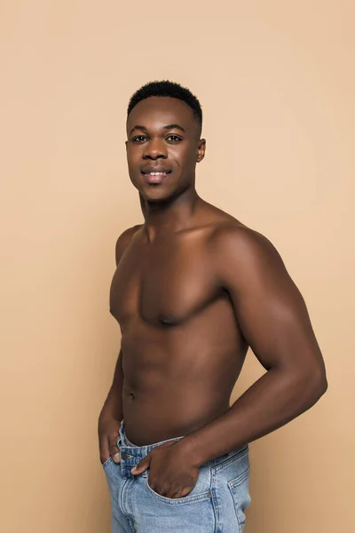 Hombre afroamericano sin camisa sonriendo mientras está de pie con las manos en bolsillos aislados en beige - foto de stock