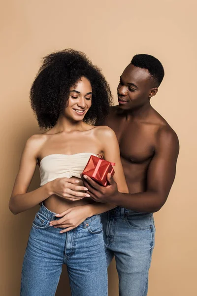 Shirtless african american man giving wrapped present to cheerful woman on beige — Stock Photo
