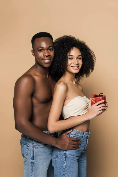 Shirtless african american man hugging happy woman with present on beige — Stock Photo