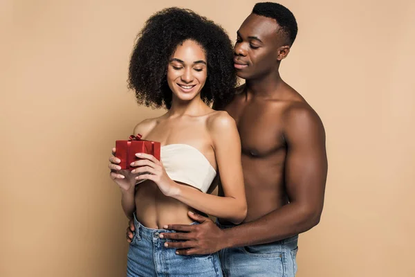 Shirtless african american man hugging cheerful woman with present on beige — Stock Photo