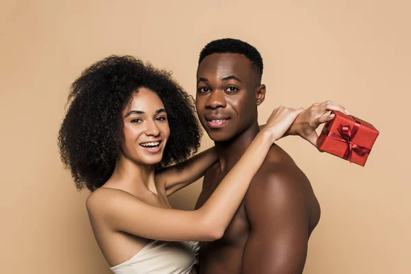 Feliz mulher afro-americana segurando presente e abraçando namorado sem camisa isolado no bege — Fotografia de Stock