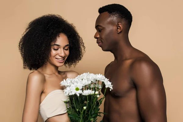 Felice donna afro-americana guardando i fiori vicino al fidanzato senza maglietta isolato sul beige — Foto stock