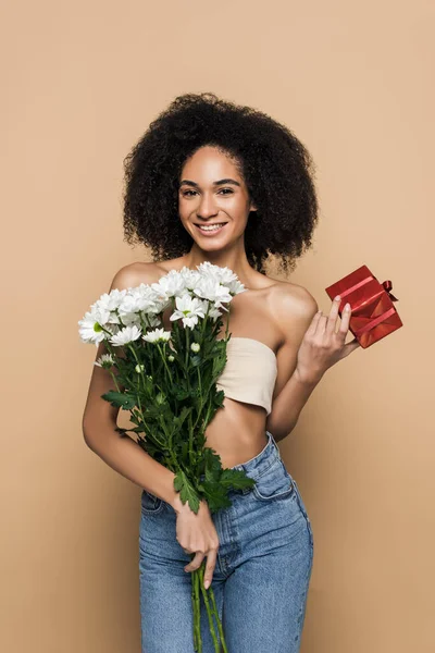 Mulher americana africana feliz com ombros nus segurando presente e flores no bege — Fotografia de Stock