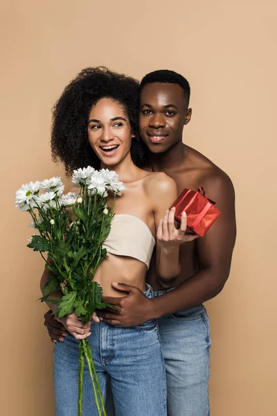 Uomo afroamericano senza maglietta che abbraccia donna riccia con fiori e presente sul beige — Foto stock