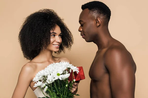 Без сорочки афроамериканський чоловік дарує квіти і подарунок щасливій жінці ізольовані на бежевому — стокове фото
