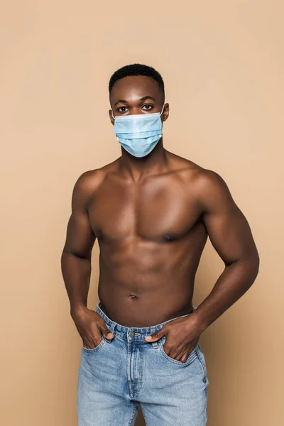 Африканский мужчина без рубашки в медицинской маске стоит с руками в карманах, изолированных на бежевом — стоковое фото