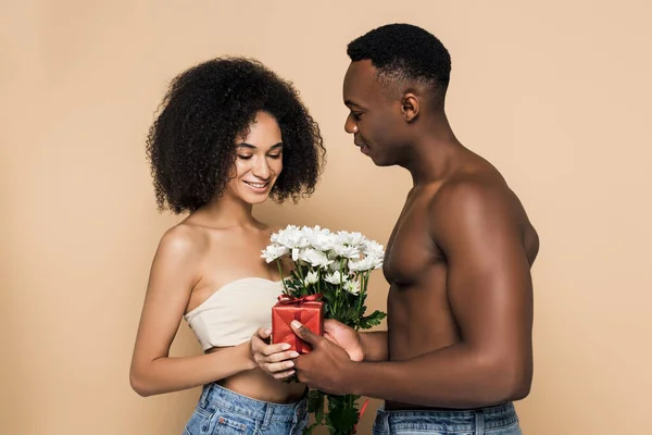 Hemdloser afrikanisch-amerikanischer Mann überreicht Blumen und Geschenke an fröhliche Frau isoliert auf beige — Stockfoto