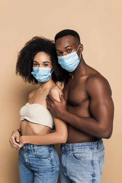 Young african american man in medical mask looking at camera while hugging girlfriend on beige — Stock Photo