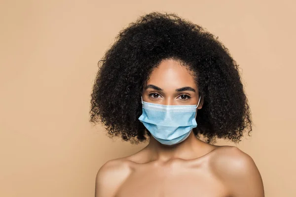 Femme afro-américaine bouclée en masque médical avec les épaules nues regardant la caméra isolée sur beige — Photo de stock