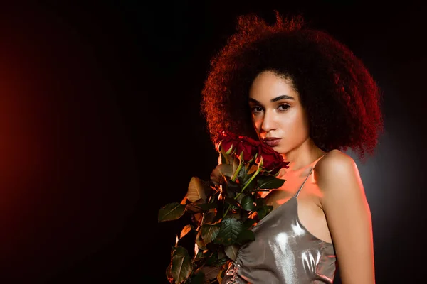 Femme afro-américaine bouclée en robe tenant des roses rouges sur noir — Photo de stock